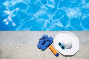 sandals, sunscreen, sunglasses, and a hat laying next to a swimming pool